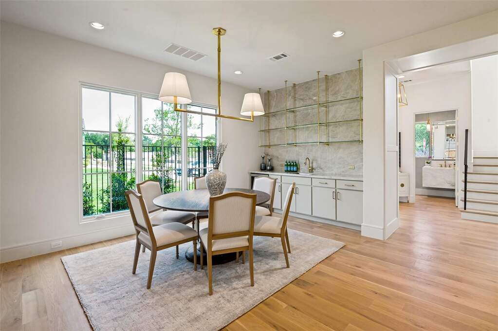 Open-concept dining area featuring light wood-finish flooring, a contemporary linear chandelier, and a built-in wet bar with a stone backsplash and glass shelving