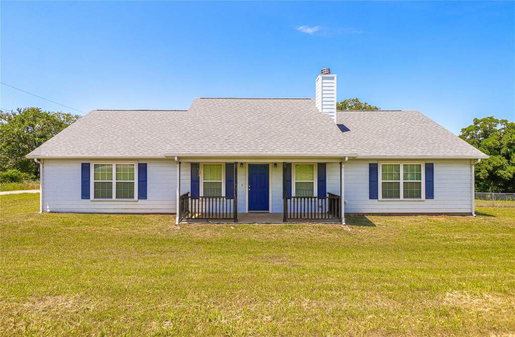 Ranch-style home with white lap siding, blue shutters, covered front porch with wood railing, and composition shingle roof. Chimney and mature trees visible on a spacious lot.