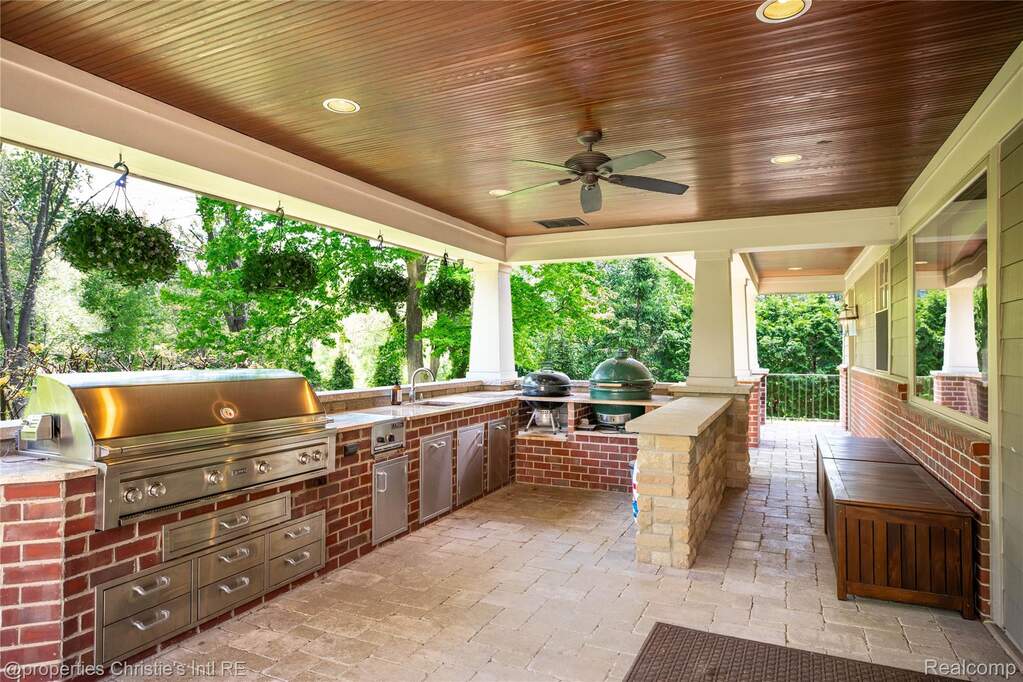 Covered outdoor kitchen featuring a built-in stainless steel grill, integrated sink, brick and stone base, and wood-finish ceiling with recessed lighting and a ceiling fan