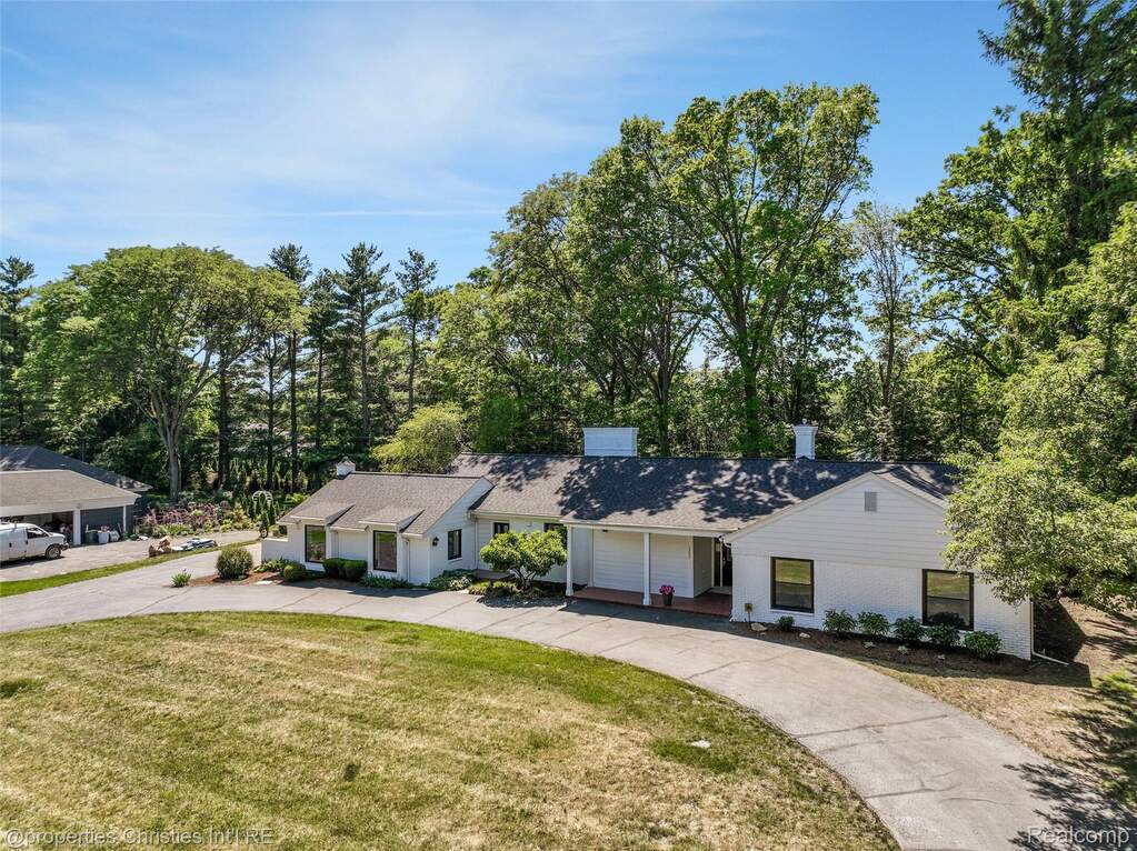 Expansive property featuring a white exterior, a long paved driveway, a covered entry porch, and mature surrounding trees