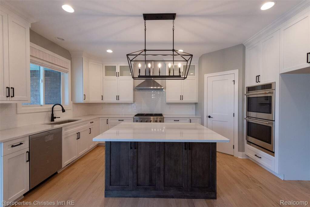 Spacious kitchen featuring a central island with wood-finish cabinetry, light-toned wood-finish flooring, white upper and lower cabinetry, stainless steel double ovens, and a geometric chande