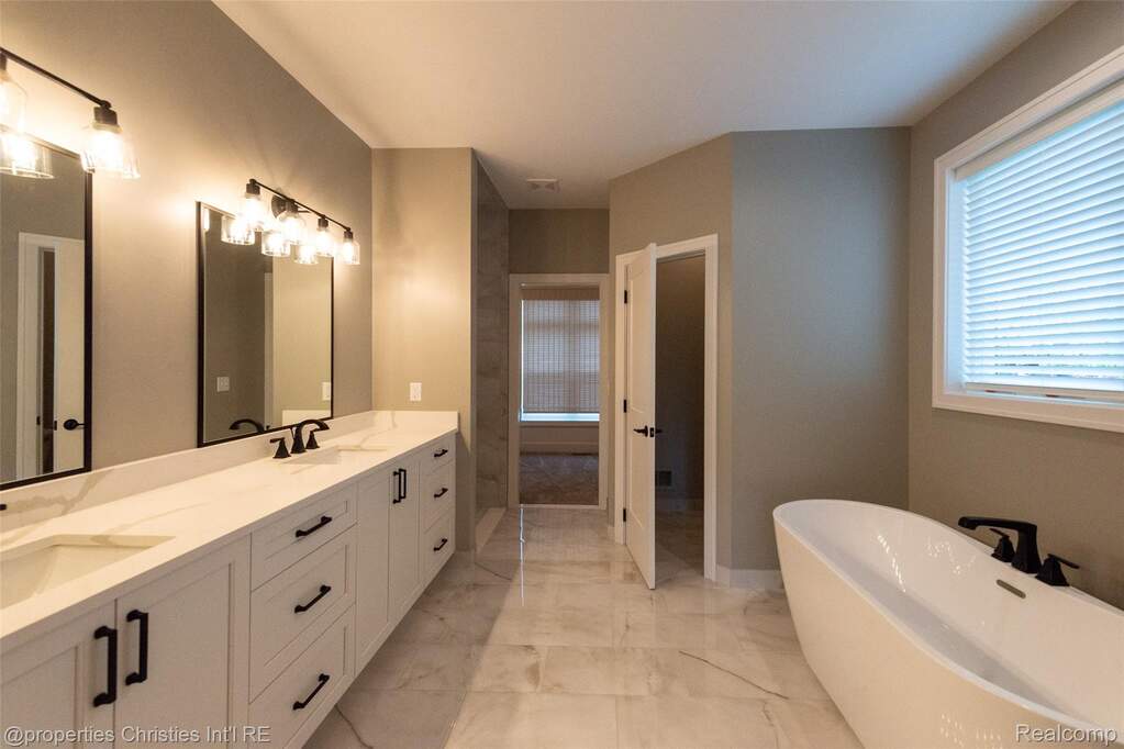 Spacious bathroom featuring a double vanity with stone countertops, an oval freestanding tub, large format marble-look floor tiles, and abundant natural light from the window
