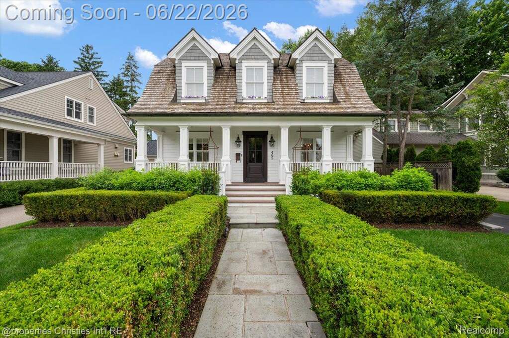 Dormered Gambrel-style home featuring a covered front porch with white pillars, a dark wood entry door, and a stone walkway framed by manicured hedges