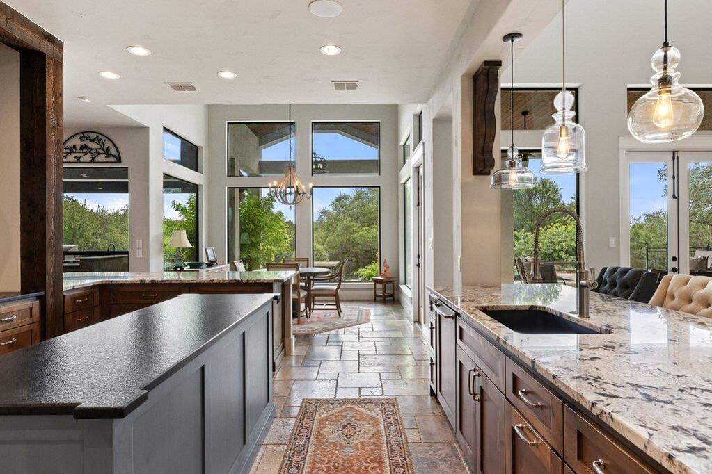 An airy kitchen features rich wood cabinetry, stone flooring, and high ceilings, all illuminated by recessed and pendant lighting.