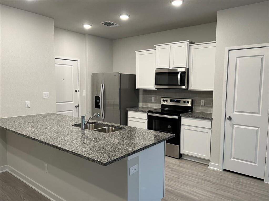 Well-appointed kitchen featuring an island with a dual-basin stainless steel sink, granite countertops, stainless steel appliances, white cabinetry, and wood-finish flooring