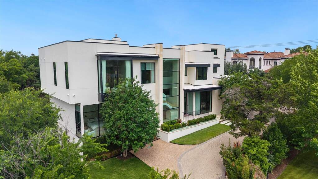 Modern architectural residence featuring a prominent glass-enclosed staircase, expansive windows, clean white stucco exterior, a paver driveway, and manicured landscaping