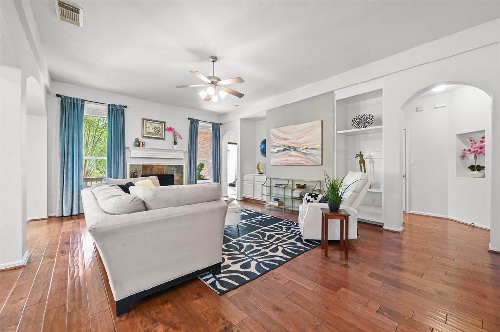 Spacious living area featuring wood-finish flooring, a fireplace with a stone surround and mantel, a ceiling fan, built-in shelving, and arched doorways