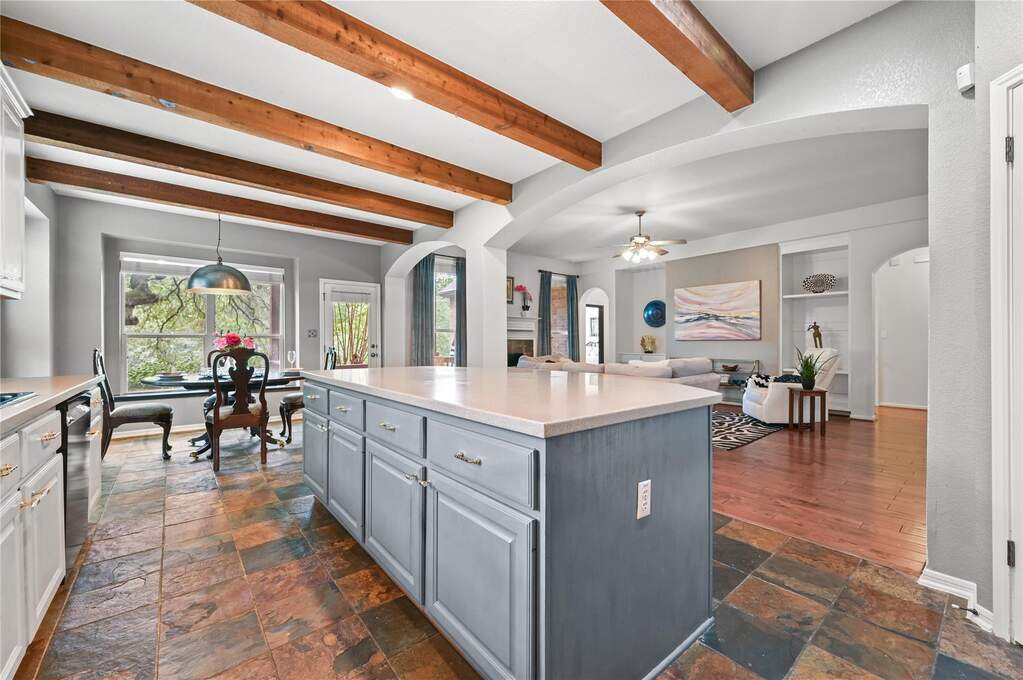 Spacious kitchen featuring a large central island with light-toned countertop, wood-finish ceiling beams, and multi-toned tile flooring