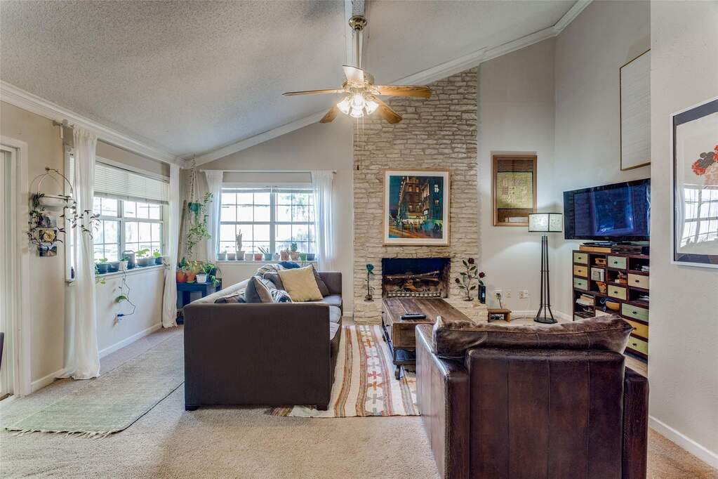 Living room featuring a textured ceiling, a stone fireplace, ornamental molding, light carpet, and ceiling fan